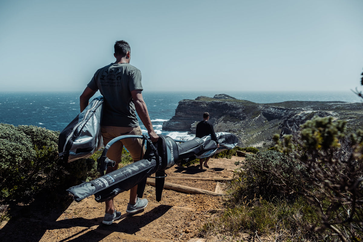 Twee windsurfers lopen met hun materiaal over een kustpad naar zee, met een North Ultimate RDM 370 in de mastzak