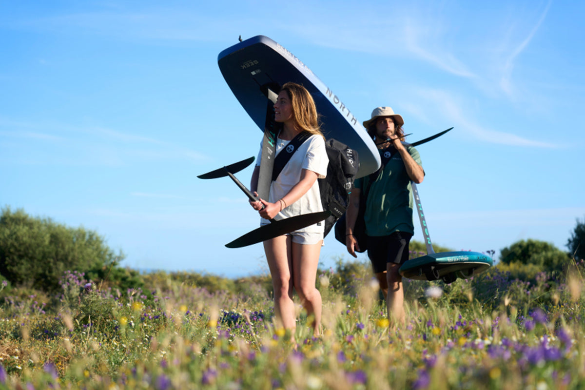 Twee wingfoilers lopen door een bloemenveld met de North Seek Foil Board 2025 en foils naar het water