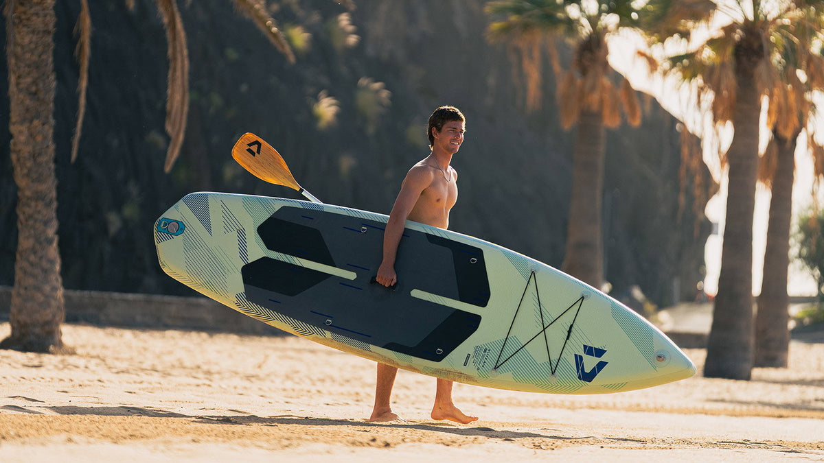 Duotone Blitz Air SLS opblaasbare touring-SUP gedragen over een zonnig strand met palmbomen