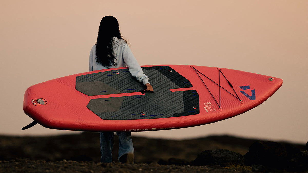 Duotone Grace Air SLS opblaasbare SUP in koraalrood, onder de arm meegedragen aan de kust