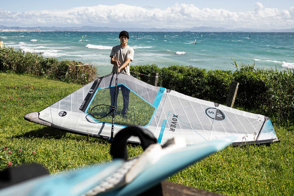 Windsurfer tuigt het North X-Over GEN2-zeil op, op het gras boven het strand
